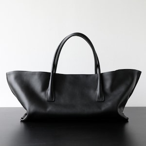 May include: A black leather tote bag with two curved handles. The bag has a wide, open top and a simple, minimalist design. The bag is sitting on a black surface against a white background.