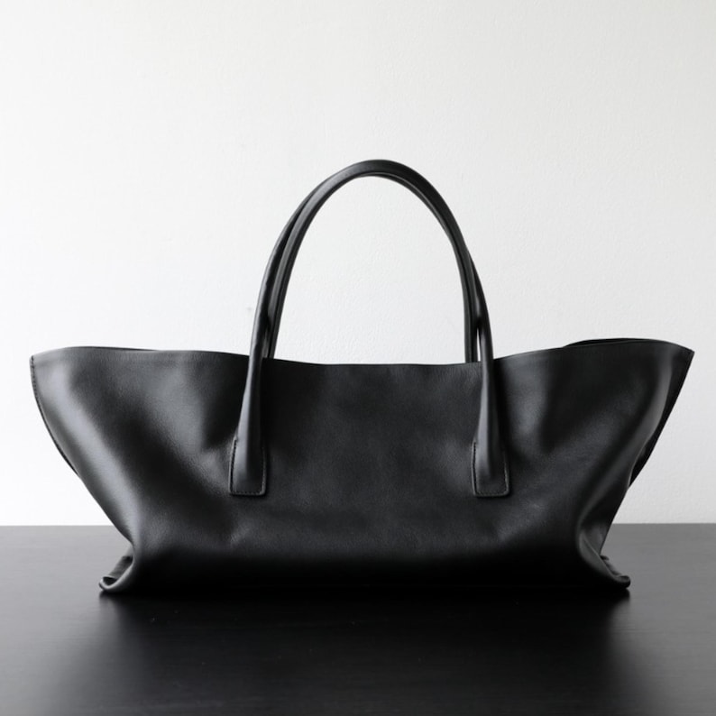 May include: A black leather tote bag with two curved handles. The bag has a wide, open top and a simple, minimalist design. The bag is sitting on a black surface against a white background.