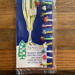 May include: A set of twelve small plastic glass markers attached to a blue cardboard backing. The packaging features a yellow champagne flute illustration and text reading GLASS-MARKERS TO IDENTIFY YOUR DRINKS and DECORATE WITH GREETING CARDS.