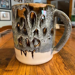 May include: A ceramic mug featuring a reactive glaze with a metallic bronze top section that drips down into a textured grey and white speckled middle, transitioning to a solid cream base. The letters NY are clearly visible in black near the bottom.
