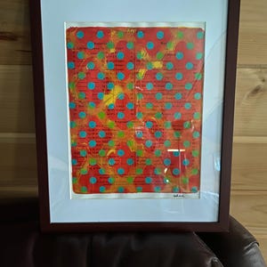 Op de afbeelding: Ingelijst abstract kunstwerk met een rode en gele achtergrond en turquoise stippen. Het kunstwerk zit in een donkerbruine houten lijst met een witte passe-partout. Het kunstwerk lijkt op een pagina tekst te staan.