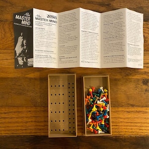 May include: A Mini Master Mind game set featuring a folded instruction manual with black text on white paper, a rectangular brown plastic game board with grid holes, and a separate brown plastic tray filled with small, multi-colored plastic pegs.