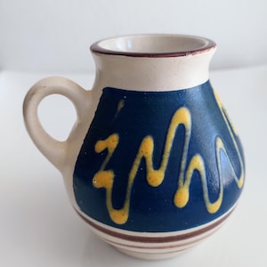 May include: A small ceramic jug with a rounded body and a single handle. The base is off-white with brown horizontal stripes. The upper section features a deep blue glaze decorated with a vibrant yellow, wavy, hand-painted line pattern.