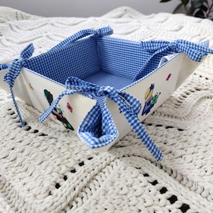 May include: A fabric bread basket with a blue and white gingham interior and trim. The exterior is off-white with illustrations of a person gardening. Blue gingham bows secure the corners. The basket sits on a white, textured surface.