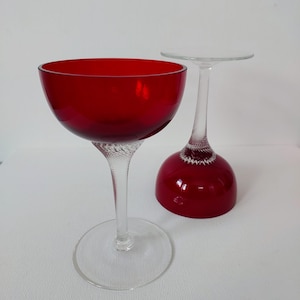 May include: Two stemmed glasses with deep red bowls and clear glass stems featuring a delicate white latticino twist pattern. One glass stands upright on a circular base, while the second glass is inverted, resting on its rim with the base facing up.