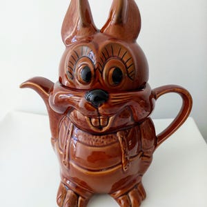 May include: A glossy brown ceramic teapot shaped like a cartoon squirrel. The teapot features large eyes with black lashes, a black nose, visible front teeth, and a handle on the right side. The lid forms the top of the squirrel's head and ears.