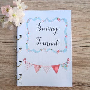 Może przedstawiać: Biały dziennik szycia z niebieskim i różowym wzorem kwiatowym. Okładka zawiera tekst "Sewing Journal" w ozdobnym kroju pisma.