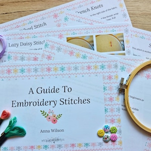 Op de afbeelding: Een gedrukte gids getiteld A Guide To Embroidery Stitches door Anna Wilson, met kleurrijke bloemenranden, instructieve tekst en diagrammen. Bevat een houten borduurring, diverse kleine ronde knopen en groen borduurgaren.