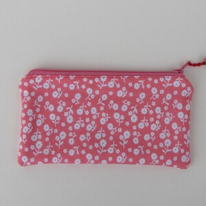 May include: A pink zippered pouch with a white floral pattern. The pouch has a pink zipper and a small green bead on the zipper pull.