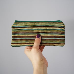 Puede incluir: Un estuche para l&aacute;pices de tela a rayas con una cremallera verde. El estuche tiene rayas marrones, verdes y azules.
