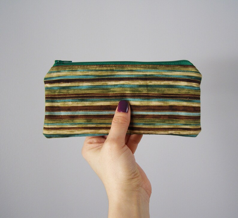 Puede incluir: Un estuche para l&aacute;pices de tela a rayas con una cremallera verde. El estuche tiene rayas marrones, verdes y azules.