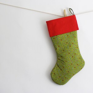 Op de afbeelding: Een kerstsok met een rode boord en een groene body versierd met kleine, kleurrijke bloempatronen. De sok hangt aan een waslijn met een houten knijper. De achtergrond is effen wit.