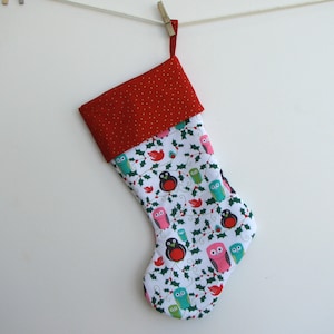 May include: A Christmas stocking with a red cuff and a white body. The cuff has a gold polka dot pattern. The body is patterned with colorful owls, birds, and holly on a white background. The stocking is hanging from a clothesline.