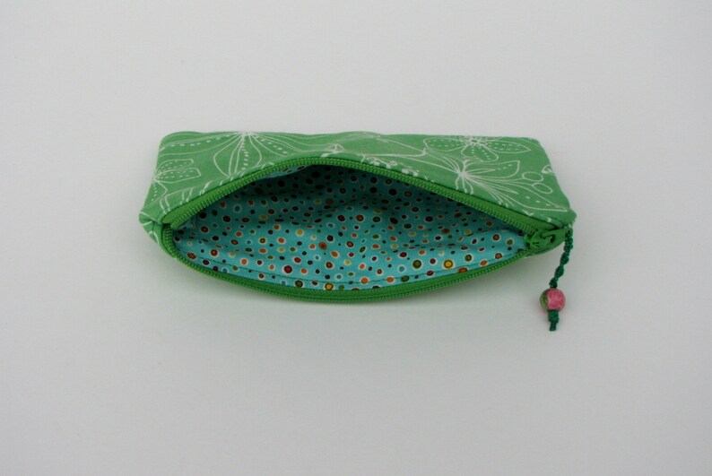 May include: A green zippered pouch with a white floral pattern on the outside and a turquoise polka dot lining on the inside. The pouch has a pink and green tassel.