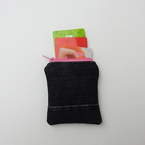 May include: A small, black denim coin purse with a pink zipper and trim. The purse is open, revealing several colorful cards inside. The purse is approximately 4 inches tall and 3 inches wide.
