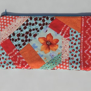 以下が含まれることがあります： 赤、オレンジ、青の配色でパッチワークされたジッパー付きポーチ。ポーチは、水玉模様、花柄、幾何学模様など、さまざまなパターンを持つ異なる生地で作られています。ポーチの中央には、大きなオレンジ色の花が飾られています。