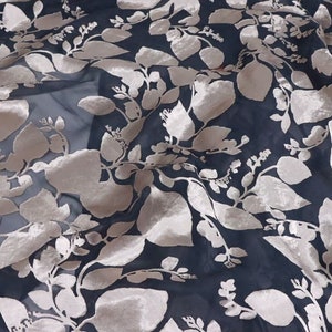 Può includere: Un tessuto nero trasparente con un delicato motivo floreale di colore beige chiaro. Il tessuto ha una leggera lucentezza e una texture morbida e fluida.