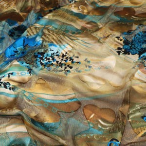 May include: A sheer fabric featuring a layered pattern of blue floral motifs, brown abstract shapes, and horizontal teal stripes. The material has a subtle metallic gold thread woven throughout, creating a shimmering texture across the surface.