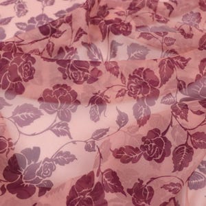 May include: A sheer, lightweight fabric in a soft peach hue, featuring a repeating pattern of dark plum-coloured roses and leaves. The material is translucent, with folds creating depth and highlighting the floral print across the surface.