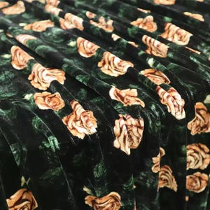 Op de afbeelding: Een donkergroene fluwelen stof met een herhalend patroon van lichtbruine en crèmekleurige rozen. Het materiaal is gedrapeerd in zachte plooien, wat de textuur van het fluweel en de bloemenprint op het oppervlak van het textiel benadrukt.