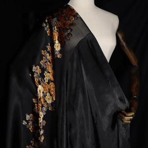 May include: A black fabric drape featuring a vertical floral pattern in shades of gold, bronze, and brown. The material has a smooth, satin-like sheen and is displayed on a mannequin, showcasing the intricate embroidery along one side of the cloth.