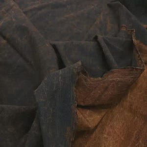 Könnte beinhalten: Ein Stoffstück mit zweifarbigem Design, bestehend aus einer dunkel marineblauen Seite und einer kontrastierenden warmbraunen Seite. Das Material weist eine stark texturierte, gesprenkelte Oberfläche mit sichtbaren Falten auf.