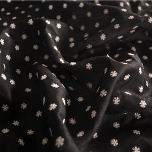 Puede incluir: Tela negra con un patrón de pequeñas formas florales blancas bordadas. La tela está drapeada, creando pliegues y sombras suaves. El patrón floral se distribuye uniformemente por todo el material, ofreciendo una apariencia delicada y texturizada.