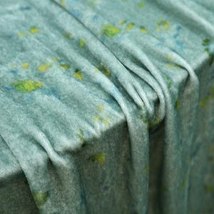Op de afbeelding: Een zachte, lichtblauwe fluwelen stof met een gevlekt patroon van verspreide gele en donkerblauwe vlekken. Het materiaal is gedrapeerd in diepe, vloeiende plooien, wat de pluche textuur en de subtiele kleurvariaties op het oppervlak benadrukt.