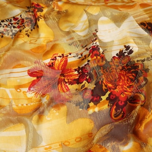 Op de afbeelding: Een gele stof met een bloemenpatroon. De stof is doorschijnend, bijna transparant, met een subtiele glans. De bloemen zijn rood, oranje en bruin en zijn in een willekeurig patroon gerangschikt.