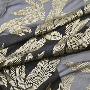May include: A sheer black fabric featuring a repeating metallic gold leaf pattern. The material is draped in soft folds, highlighting the contrast between the dark, translucent base and the textured, shimmering gold foliage design throughout.