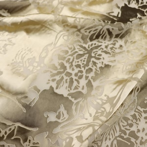 Puede incluir: Primer plano de una tela con un estampado floral. La tela tiene una base color crema con un efecto sombreado sutil en tonos grises y marrones. El diseño floral es de un color blanco roto más claro, creando un contraste delicado. La tela está doblada.