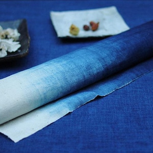 May include: A rolled piece of fabric featuring an ombre gradient effect that transitions from a light, off-white color at one end to a deep, saturated indigo blue at the other. The textile has a visible woven texture and raw, unfinished edges.