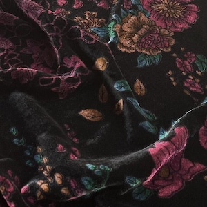 Op de afbeelding: Een zwarte fluwelen stof met een bloemenpatroon in diep magenta, brons en blauwgroen. Het materiaal heeft een zachte, pluche textuur met zichtbare plooien en draperingen die het ingewikkelde botanische ontwerp van bloemen en bladeren benadrukken.