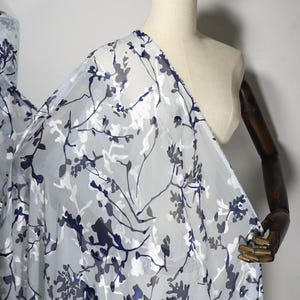 May include: A sheer, lightweight fabric featuring a printed pattern of dark navy blue and muted grey floral branches and leaves scattered across a translucent white background, draped over a mannequin to display the semi-transparent texture.