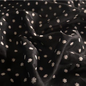 May include: A black fabric featuring a repeating pattern of small, white embroidered floral motifs. The material is draped in soft folds, highlighting the texture of the embroidery and the smooth, dark surface of the textile throughout the piece.