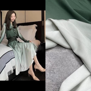 Peut inclure: Un tissu de soie à dégradé vert et blanc. Le tissu est lisse et a un léger brillant. Le tissu est drapé sur une surface grise.