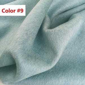 May include: A light blue fabric with a soft, fuzzy, and dense texture. The material is draped in folds, creating shadows and highlights across the surface. A white rectangular label in the upper left corner displays the text Color #9.