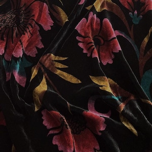Op de afbeelding: Een zwarte fluwelen stof met een grootschalig bloemenpatroon. De bloemen zijn uitgevoerd in tinten diep rood, magenta en gedempt goud, met donkergroene bladeren als accent. Het materiaal heeft een zachte, lichtreflecterende textuur met plooien.