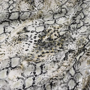 May include: A lightweight fabric featuring a layered pattern of black, white, and muted gold tones. The design combines a geometric snakeskin print with an overlaid floral motif, creating a textured visual effect across the folded material.