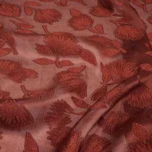 May include: A sheer, translucent fabric in a deep terracotta red hue, featuring an all-over embroidered floral pattern. The flowers are rendered in a darker, saturated red thread, creating a textured, raised appearance against the semi-transparent base.