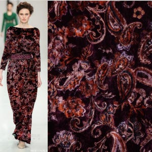 May include: A long-sleeved floor-length dress featuring a dark velvet fabric with an intricate paisley and floral pattern in shades of deep burgundy, rust orange, and muted purple, accented by a matching wide belt with a geometric buckle.
