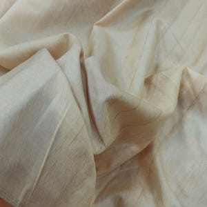 Puede incluir: Primer plano de una tela beige con un patrón sutil y texturizado. La tela está doblada, creando sombras y reflejos suaves. El material parece ligero y fluido, con un aspecto natural y orgánico.