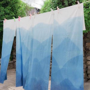 Peut inclure: Trois panneaux de tissu rectangulaires suspendus à une corde à linge avec des pinces en plastique colorées. Le tissu présente une section supérieure blanche qui se transforme en un motif de montagnes en dégradé bleu vers le bas.