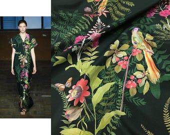 May include: A floor-length dark green dress featuring a vibrant botanical print with colorful birds, butterflies, red berries, and various green fern and leaf patterns. The garment has short sleeves and a V-neckline, showcasing a detailed nature-inspired design.