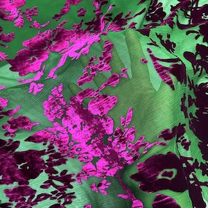 Op de afbeelding: Een transparante, gekreukte groene stof met een verhoogd, fluweelachtig bloemenpatroon in levendig magenta en dieppaars. Het materiaal is doorschijnend, waardoor licht door het delicate, getextureerde weefsel van de groene basisstof valt.