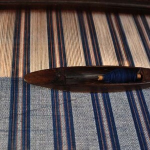 May include: A wooden loom with a blue and white striped fabric being woven. A wooden shuttle with blue thread is resting on the loom.
