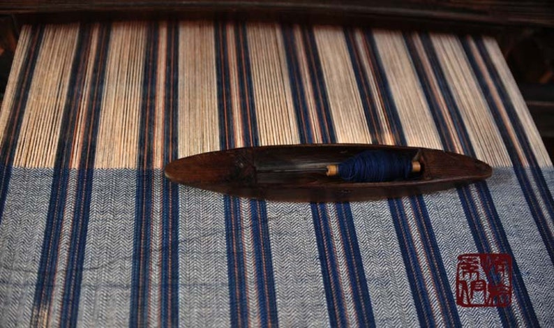 May include: A wooden loom with a blue and white striped fabric being woven. A wooden shuttle with blue thread is resting on the loom.