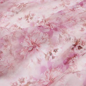Pode incluir: Close-up de um tecido transparente com um padrão floral. O tecido apresenta grandes flores rosa macias com detalhes em branco e rosa claro. As flores estão espalhadas pelo tecido, criando um design delicado e romântico.