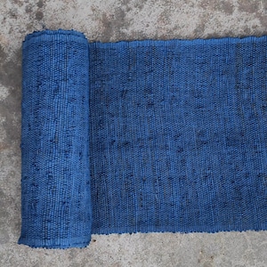 Pode incluir: Um tapete azul texturizado enrolado, com um padrão tecido. O tapete é de um tom azul vibrante, com manchas mais escuras em toda a trama. O tapete está desenrolado, revelando mais da superfície texturizada.