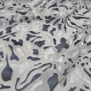 May include: A sheer, lightweight fabric featuring an abstract pattern of dark charcoal gray and opaque white organic shapes. The material has a semi-transparent quality with a textured surface, displaying a repeating irregular camouflage-like design.